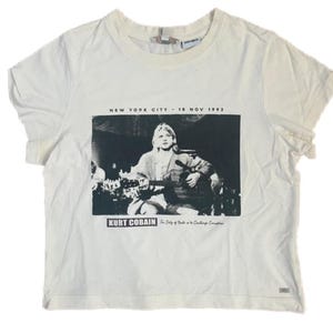 Può includere: Una t-shirt a maniche corte color bianco sporco con una grafica in bianco e nero di Kurt Cobain che suona la chitarra. Il testo NEW YORK CITY - 18 NOV 1993, KURT COBAIN e The Duty of Youth is to Challenge Corruption è stampato sul davanti.