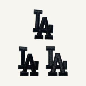May include: Three black embroidered patches featuring the interlocking LA logo. Each patch displays the capital letters L and A in a bold, block-style font with a textured finish, arranged with one patch centered above two others in a triangular formation.