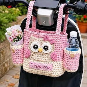 Peut inclure: Un sac au crochet rose et beige en forme de hibou avec deux anses, orné d'un petit nœud rose, de grands yeux et d'une étiquette rectangulaire portant le nom Vanessa. Le sac possède deux poches latérales, l'une contenant des fleurs et l'autre une bouteille.