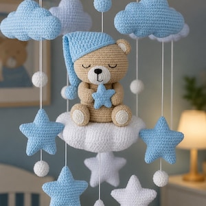 May include: A crochet baby mobile featuring a central tan bear wearing a blue nightcap and holding a blue star. The bear sits on a white cloud, surrounded by hanging blue and white crocheted clouds, stars, and small spheres suspended from a circular frame.