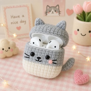 Può includere: Una custodia all'uncinetto grigia e crema per auricolari wireless, a forma di gatto con orecchie a punta, una piccola coda, occhi a perla neri, naso ricamato e guance rosa. La custodia è aperta e mostra gli auricolari bianchi all'interno.