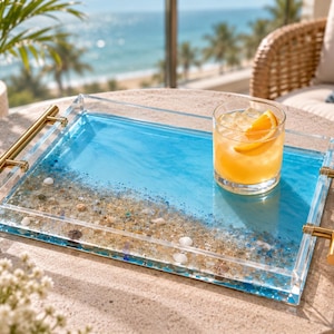 May include: A rectangular clear acrylic serving tray featuring a decorative resin inlay that mimics a beach scene with blue water and sandy shore with small shells. The tray has two gold-toned metal handles attached to the sides.