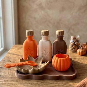 May include: A set of three silicone bottles in orange, beige, and brown, each featuring a different embossed design of a pumpkin, leaf, or acorn. The set includes a leaf-shaped bowl, a matching orange spoon, and a small orange pumpkin-shaped cup.
