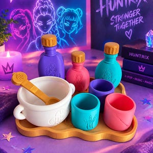 May include: A Huntrix toy set featuring a white cauldron with a gold spoon, three colored bottles with gold caps, and three matching cups on a gold tray. The items feature embossed character graphics and symbols in purple, pink, and teal.
