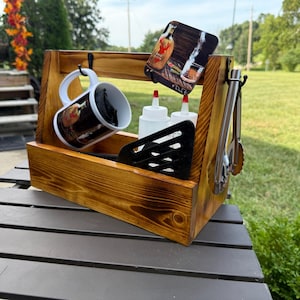 May include: A rectangular wooden caddy with a dark stained finish, featuring a handle and metal hooks. It holds a white ceramic mug with a graphic print, two clear plastic squeeze bottles, a black slotted spatula, and a pair of metal tongs.