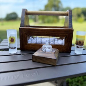 May include: A dark stained wooden caddy with a wire mesh front, a wooden handle, and a small matching wooden base holding a clear tea light. Two frosted glass shot glasses feature sunflower graphics and the word Kansas printed in black text.
