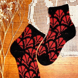 May include: A pair of ankle-length knit socks featuring a repeating geometric pattern in bold red and black. The socks have solid black cuffs, heels, and toes, with the red and black design covering the main body of the foot and the ankle section.