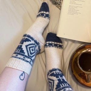 May include: A pair of cream-colored knit socks featuring a dark blue geometric pattern around the ankle and a solid dark blue toe section. The socks have a ribbed cuff and a detailed, repeating motif that includes angular shapes and small star accents.