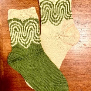 May include: A pair of knitted socks featuring a two-tone color scheme of cream and olive green. One sock has a solid olive green foot with a cream and green wavy pattern on the cuff, while the other has a solid cream foot with a matching pattern.