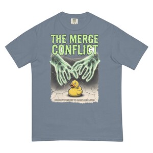 May include: A slate blue short-sleeve t-shirt featuring a graphic print with the text THE MERGE CONFLICT in dripping lime green letters, two glowing digital hands reaching down toward a small yellow rubber duck, and the text NOBODY PUSHES TO MAIN AND LIVES.