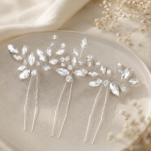 Può includere: Tre forcine color argento con un design a forma di ramo, decorate con cristalli trasparenti sfaccettati. Ogni forcina presenta una base a due punte e una parte superiore decorativa composta da pietre taglio marquise e rotondo.
