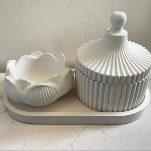May include: A set of three white matte objects consisting of an oval tray, a small bowl with a scalloped petal-like rim, and a lidded jar with a ribbed texture and a spherical knob handle, all arranged together as a cohesive decorative collection.