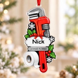 May include: A hanging ornament featuring a red pipe wrench, a roll of white toilet paper, green holly leaves with red berries, and white snowflakes. A white label displays the name Nick and the text LICENSED PLUMBER in black font.