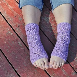 Pode incluir: Um par de meias de tricô lavanda com um padrão de renda. As meias estão sendo usadas nos pés de uma pessoa e estão apoiadas em um piso de madeira.