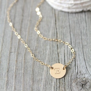 Op de afbeelding: Een delicate goudkleurige ketting met een kleine, ronde hanger. De hanger is gegraveerd met de letter "E". De ketting heeft een fijne ketting en de hanger is gecentreerd. De achtergrond is een verweerd houten oppervlak.