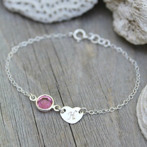 Könnte beinhalten: Ein filigranes Silberarmband mit einem rosa Edelstein und einem herzförmigen Anhänger mit dem Buchstaben "H". Das Armband liegt auf einer Holzoberfläche, im Hintergrund ein weißes korallenartiges Objekt.