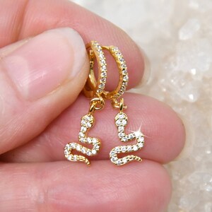 Può includere: Orecchini a cerchio color oro con un ciondolo a forma di serpente pendente. I cerchi e i ciondoli a forma di serpente sono incastonati con piccole pietre preziose trasparenti. Gli orecchini sono tenuti da una mano su uno sfondo bianco strutturato.