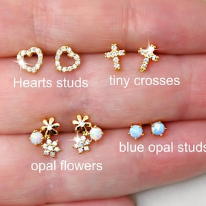 May include: A selection of gold-colored stud earrings. Styles include heart studs, tiny crosses, opal flower studs, and blue opal studs. The text "Hearts studs", "tiny crosses", "opal flowers", and "blue opal studs" are visible.