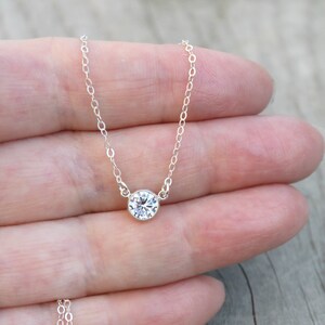 May include: A delicate silver necklace featuring a single, round, clear gemstone pendant. The pendant is set in a bezel setting and hangs from a fine silver chain. The necklace is displayed against a wooden surface.