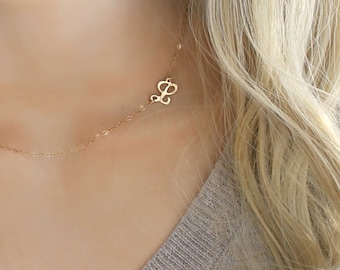 May include: A gold-colored necklace with the letter "L" pendant. The necklace is worn on a person with long, wavy blonde hair and a gray V-neck sweater. The pendant is small and delicate, adding a touch of elegance.