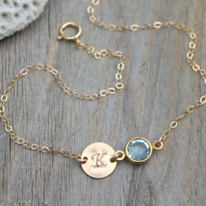 Op de afbeelding: Een delicate goudkleurige armband met een kleine ronde bedel met de gegraveerde letter "K" en een lichtblauwe edelsteen. De armband wordt getoond op een houten oppervlak met witte schelpen op de achtergrond.
