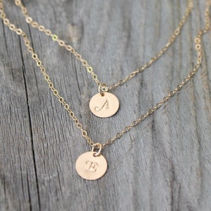 May include: Two gold-colored layered necklaces with circular pendants. The pendants are engraved with the letters "A" and "E". The necklaces are displayed on a weathered wooden surface, with greenery in the background.