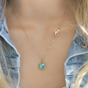 Könnte beinhalten: Eine goldfarbene Halskette mit einem türkisfarbenen Anhänger und einem kleinen Buchstaben "C"-Charm. Der Anhänger hat eine Tropfenform mit einem goldfarbenen Rand. Die Halskette wird über einem weißen Oberteil und einer Jeansjacke getragen.