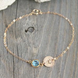 Op de afbeelding: Goudkleurige armband met een kleine, ronde, lichtblauwe edelsteen en een cirkelvormige bedel met de letter "C". De delicate kettingarmband wordt gepresenteerd op een verweerd houten oppervlak.