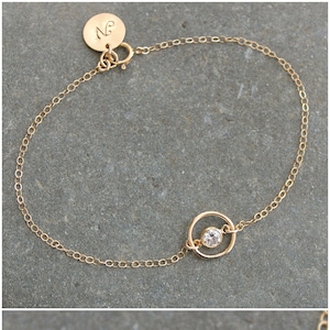 Könnte beinhalten: Zarter goldfarbener Armband mit einem kleinen, funkelnden runden Edelstein, der in einem kreisförmigen Rahmen gefasst ist. Das Armband hat eine feine Kette und einen kleinen, runden Anhänger mit den Initialen "NG".