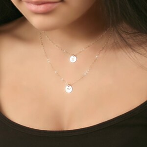 May include: Two-layered gold-colored necklace with small, round pendants. Each pendant has a delicate design. The necklace is worn against a dark top, creating a contrast that highlights the jewelry's details. The chain is delicate and the pendants are small.