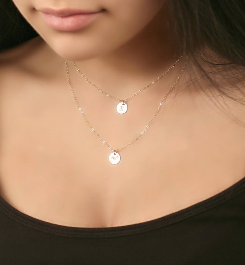 May include: Two-layered gold-colored necklace with small, round pendants. Each pendant has a delicate design. The necklace is worn against a dark top, creating a contrast that highlights the jewelry's details. The chain is delicate and the pendants are small.