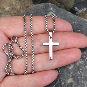 Pu&ograve; includere: Una collana con pendente a forma di croce color argento. La croce &egrave; un design semplice e lucido, sospeso a una catena abbinata. La catena ha uno stile a maglia scatolata. La collana &egrave; esposta su una mano, su uno sfondo sfocato di rocce.