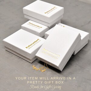 May include: Several white gift boxes with gold lettering are stacked on a gray surface. The boxes have the text "Potionumber9" and a website address. A gold necklace is partially visible. The text "YOUR ITEM WILL ARRIVE IN A PRETTY GIFT BOX Ready for Gift Giving" is at the bottom.