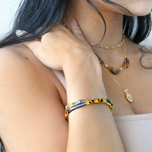 Può includere: Un braccialetto a catena dorata con motivo tartarugato e una collana a catena dorata con pendente. I gioielli sono indossati sul braccio e sul collo di una persona. Il braccialetto è largo circa 2,5 cm.