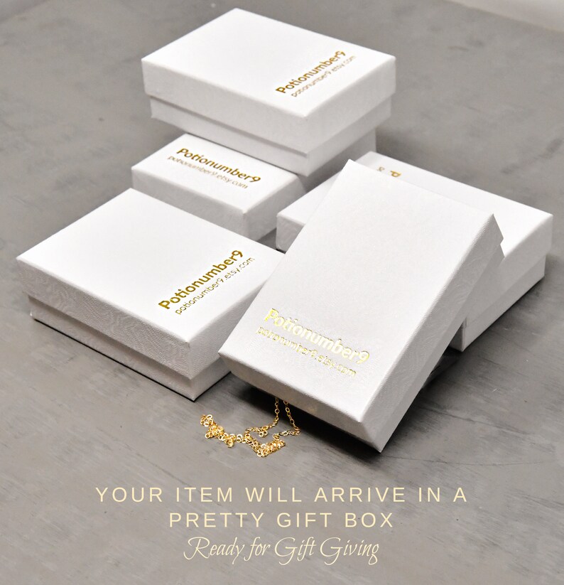 Op de afbeelding: Verschillende witte geschenkdozen met gouden letters zijn gestapeld op een grijze ondergrond. De dozen hebben de tekst "Potionumber9" en een websiteadres. Een gouden ketting is gedeeltelijk zichtbaar. De tekst "YOUR ITEM WILL ARRIVE IN A PRETTY GIFT BOX Ready for Gift Giving" staat onderaan.