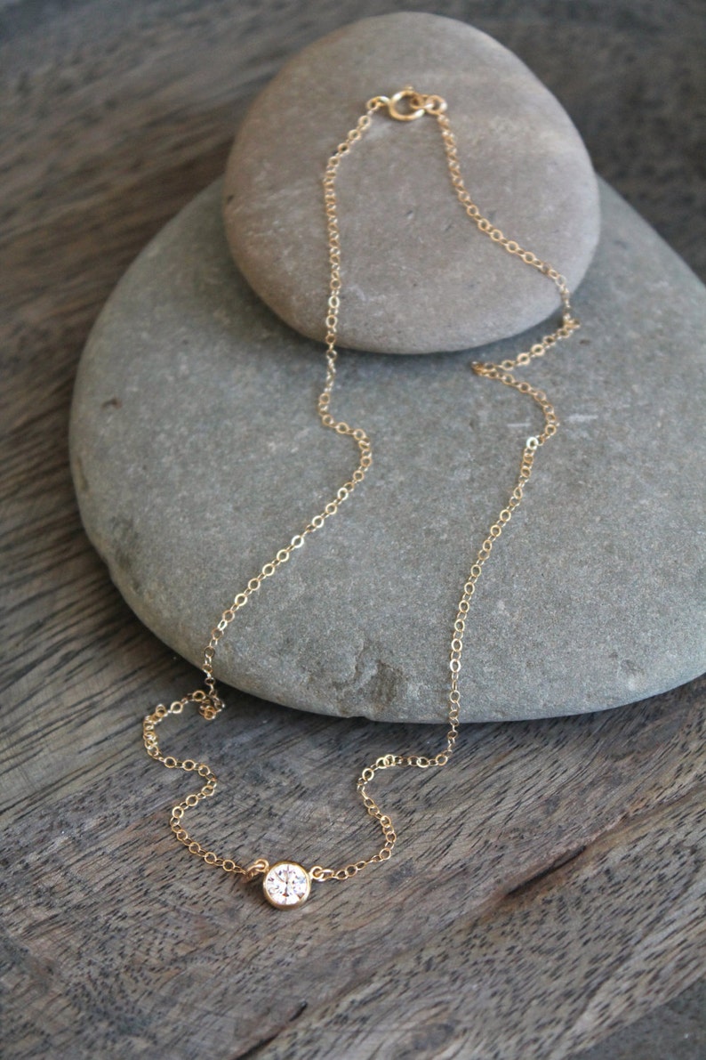 May include: A delicate gold necklace with a single, round, clear gemstone pendant. The necklace chain is fine and sits on a gray stone against a wooden surface. The clasp is visible at the top.