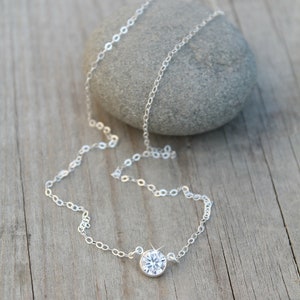May include: A delicate silver necklace with a single, round, clear gemstone pendant. The necklace chain is fine and rests on a wooden surface next to a gray stone. The gemstone sparkles, catching the light.
