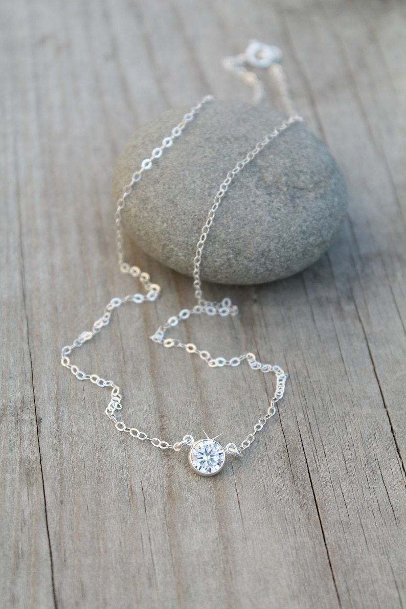 May include: A delicate silver necklace with a single, round, clear gemstone pendant. The necklace chain is fine and rests on a wooden surface next to a gray stone. The gemstone sparkles, catching the light.