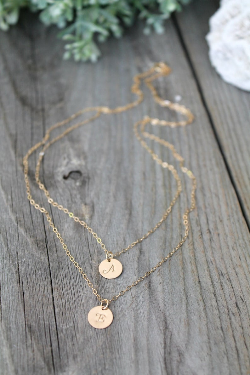 May include: Two gold-colored layered necklaces with circular pendants. The pendants are engraved with the letters "A" and "E". The necklaces are displayed on a weathered wooden surface, with greenery in the background.