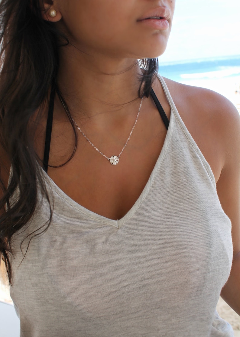 May include: A delicate silver necklace featuring a small, round pendant with intricate detailing. The pendant hangs from a fine chain. The model is wearing a light gray tank top and a black bra strap is visible.