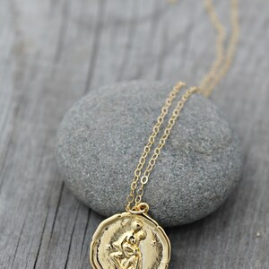 May include: A gold-colored pendant necklace featuring a detailed relief of two figures embracing. The pendant is round with a textured border and hangs from a delicate chain. The necklace rests on a gray stone against a weathered wooden surface.