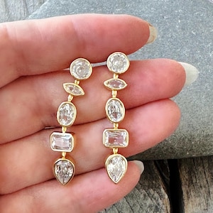 May include: Gold-tone earrings with a series of clear, faceted gemstones in various shapes: round, oval, and pear. The earrings are held up by a hand, showcasing the design and sparkle of the jewelry.