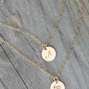 May include: Two gold-colored, layered necklaces with circular pendants. One pendant displays the letter "A", the other the letter "E". The necklaces are on a weathered, gray wooden surface.