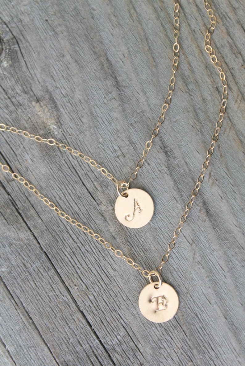 May include: Two gold-colored, layered necklaces with circular pendants. One pendant displays the letter "A", the other the letter "E". The necklaces are on a weathered, gray wooden surface.