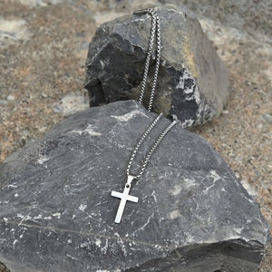 Pu&ograve; includere: Collana con pendente a croce color argento e catena a scatola. La croce ha un design semplice. La collana &egrave; esposta su una roccia grigia su uno sfondo strutturato.