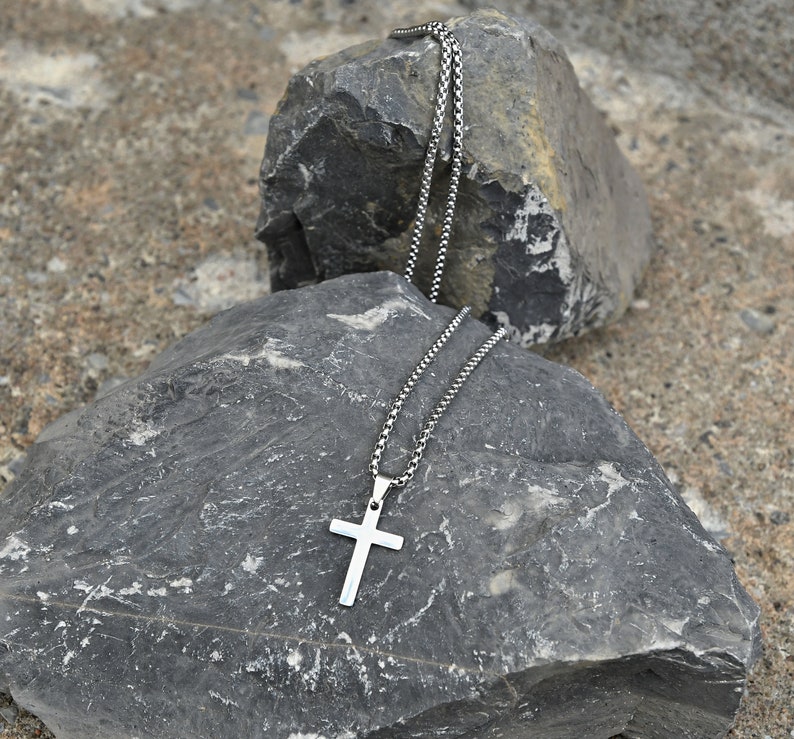 Pu&ograve; includere: Collana con pendente a croce color argento e catena a scatola. La croce ha un design semplice. La collana &egrave; esposta su una roccia grigia su uno sfondo strutturato.