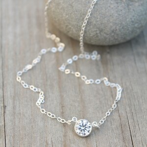 May include: A delicate silver necklace with a single, round, clear gemstone pendant. The chain is made of small, linked circles. The necklace is displayed on a wooden surface with a gray stone in the background.