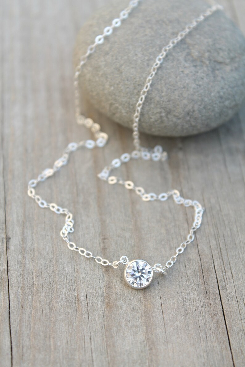 May include: A delicate silver necklace with a single, round, clear gemstone pendant. The chain is made of small, linked circles. The necklace is displayed on a wooden surface with a gray stone in the background.