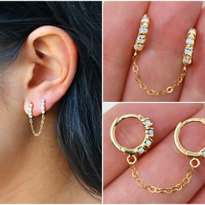 May include: Gold double hoop earrings connected by a fine chain. Each earring features a row of small, iridescent opal-like stones. The earrings are shown on an ear and also displayed separately, highlighting the design and details.