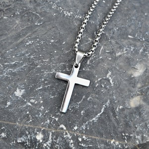 Pu&ograve; includere: Collana con pendente a croce color argento. La croce ha un design semplice e lucido. La catena &egrave; a maglie. La collana &egrave; esposta su uno sfondo grigio scuro strutturato. Simbolo religioso.